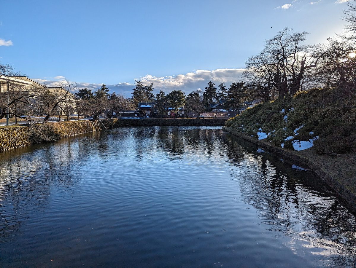 米沢城址・松が岬公園を囲む水堀と土手の残雪、青空の映る水面